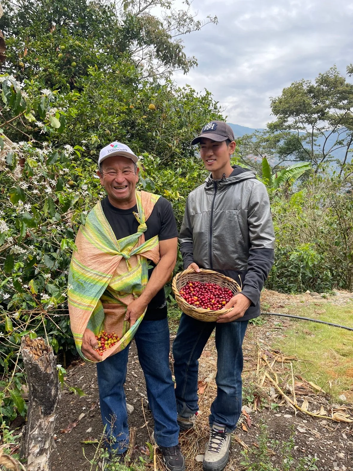 コロンビアの農園で、収穫したコーヒーチェリーの籠を抱える生産者とSENNA。背後にコーヒーの花